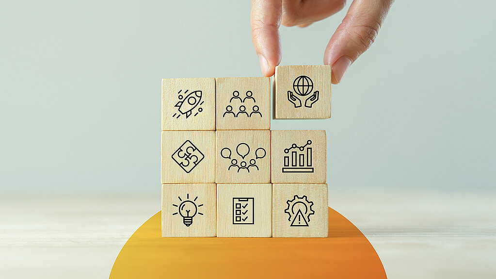 Eine Hand setzt den entscheidenden Baustein in ein Raster aus Holzw&uuml;rfeln ein, die verschiedene Erfolgsfaktoren f&uuml;r Organisationsentwicklung symbolisieren, wie Teamarbeit, Innovation und Strategie.