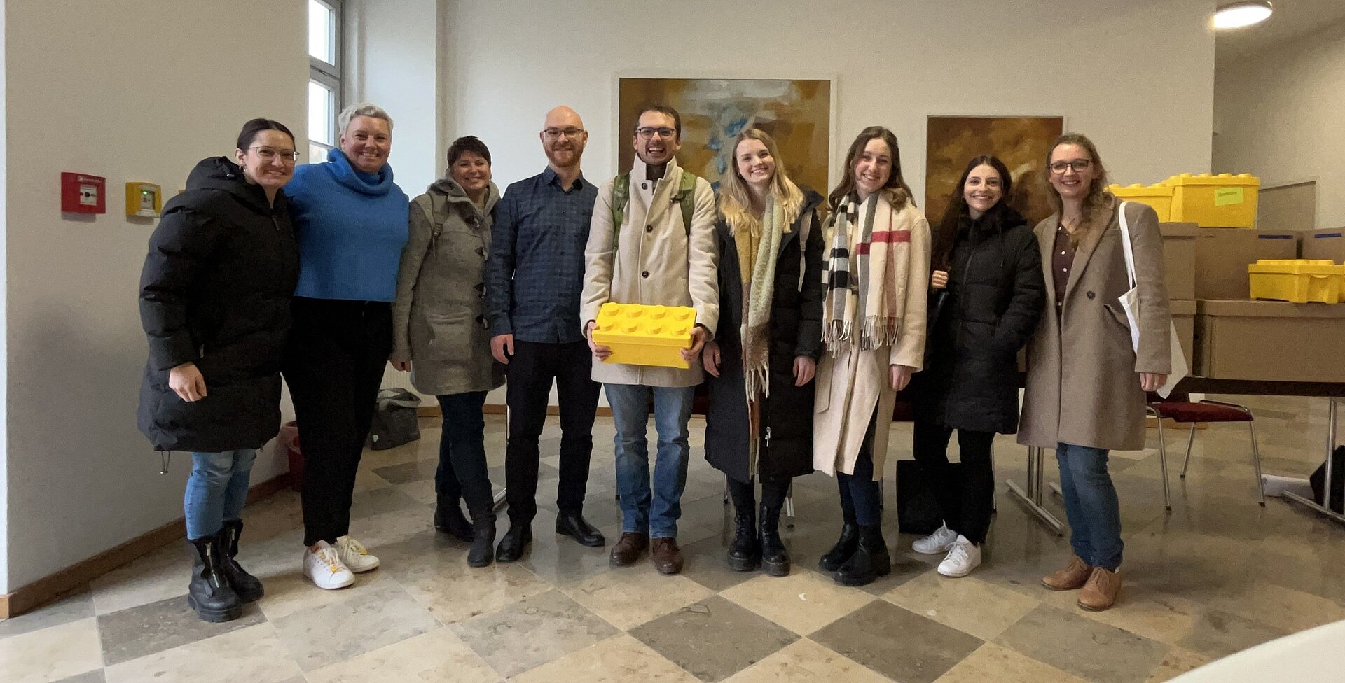 Gruppenfoto von neun LEGO® SERIOUS PLAY Trainer*innen nach einem Großgruppenworkshop. Das TEAMWILLE-Trainingsteam steht gemeinsam vor einer Präsentationsfläche und lächelt in die Kamera.