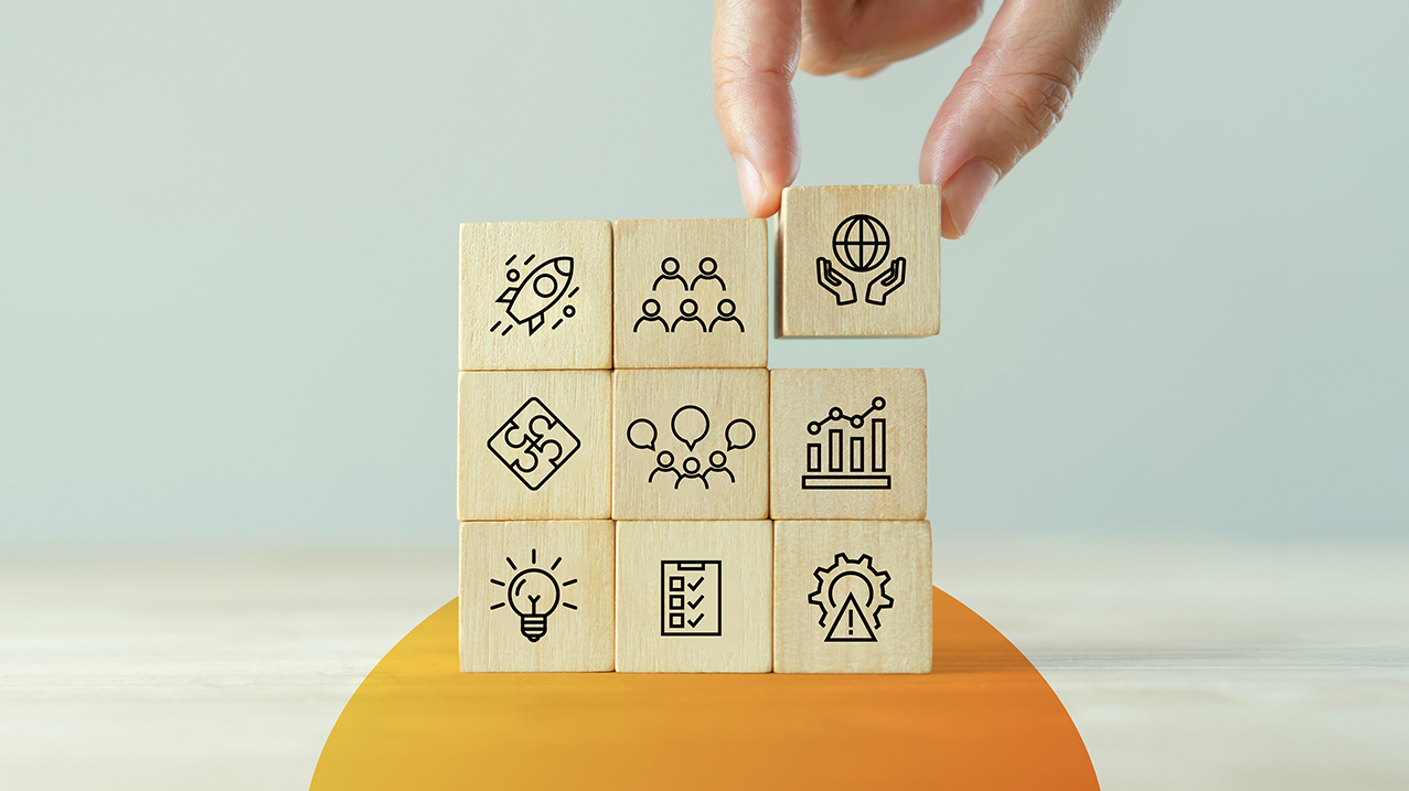 Eine Hand setzt den entscheidenden Baustein in ein Raster aus Holzw&uuml;rfeln ein, die verschiedene Erfolgsfaktoren f&uuml;r Organisationsentwicklung symbolisieren, wie Teamarbeit, Innovation und Strategie.
